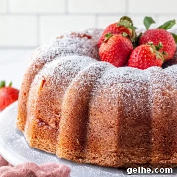 Easy Strawberry Cheesecake Bundt Cake Recipe finished on a white cake stand with powdered sugar dusting and fresh strawberries arranged on a pink linen.