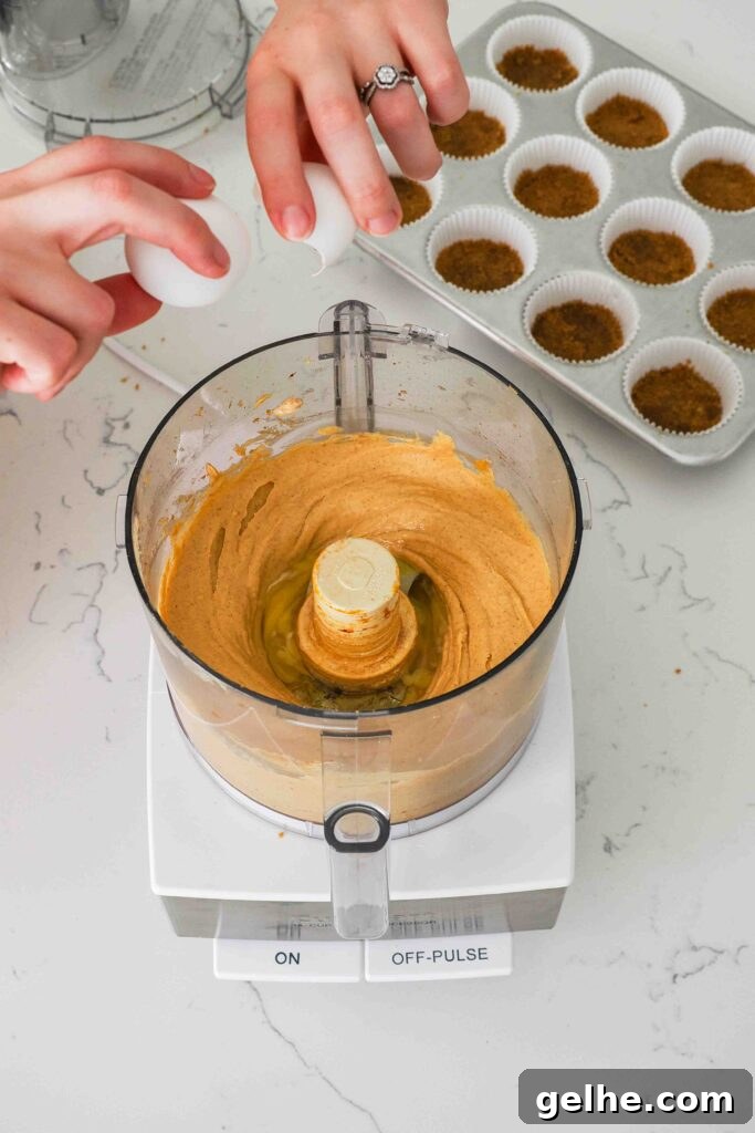 Mini Pumpkin Cheesecake Bites: Creamy Fall Dessert Recipe 9 Two hands crack an egg into a food processor bowl with the pumpkin cheesecake.