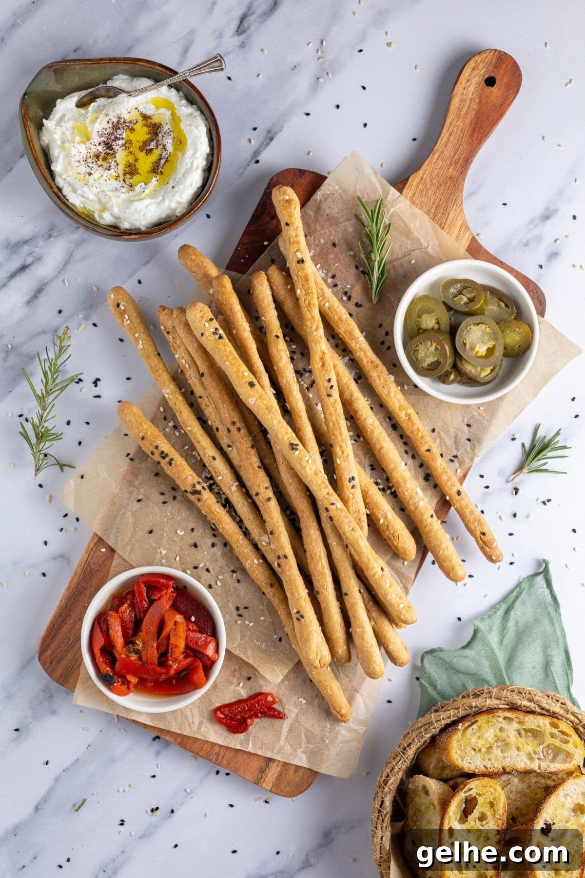 Crispy Sourdough Grissini Breadsticks for Snacking and Dipping 2 A flat lay image of crunchy sourdough breadsticks displayed on a rectangle serving board. There are some roasted red peppers and jalapenos on the board, as well as some whipped feta dip to the right. The board is garnished with fresh rosemary and there are some sourdough crostini peaking from the right corner of the image.