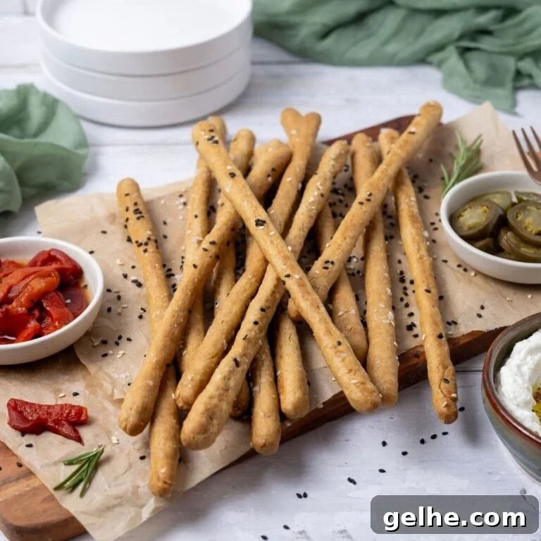 A close up photo of sourdough grissini garnished with black and white sesame seeds. The crunchy sourdough breadsticks are piled on a board surrounded by some antipasto type food.