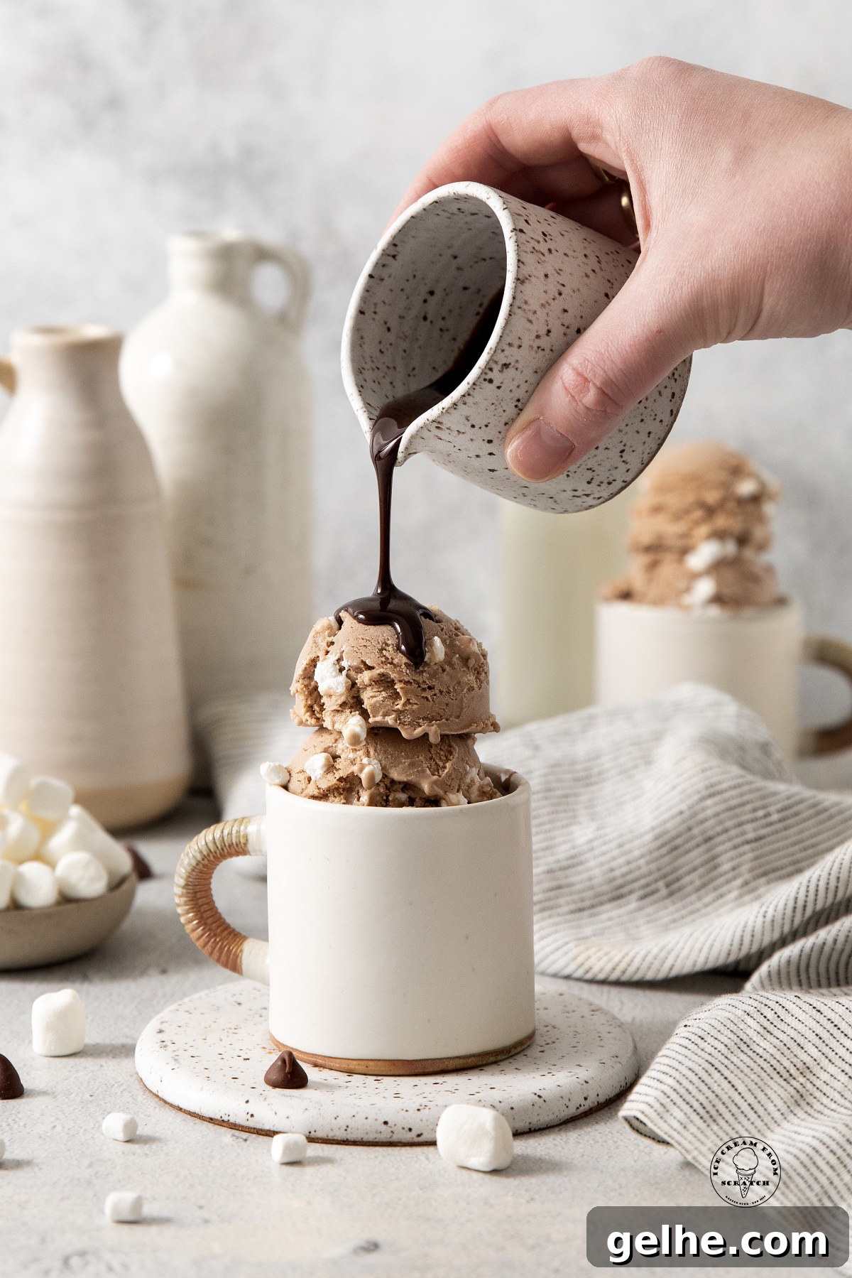 Homemade Hot Chocolate Ice Cream Recipe for Rich, Chocolatey Flavor 4 a hand pouring hot fudge from a small white and brown speckled pitcher over a mug filled with hot chocolate ice cream.