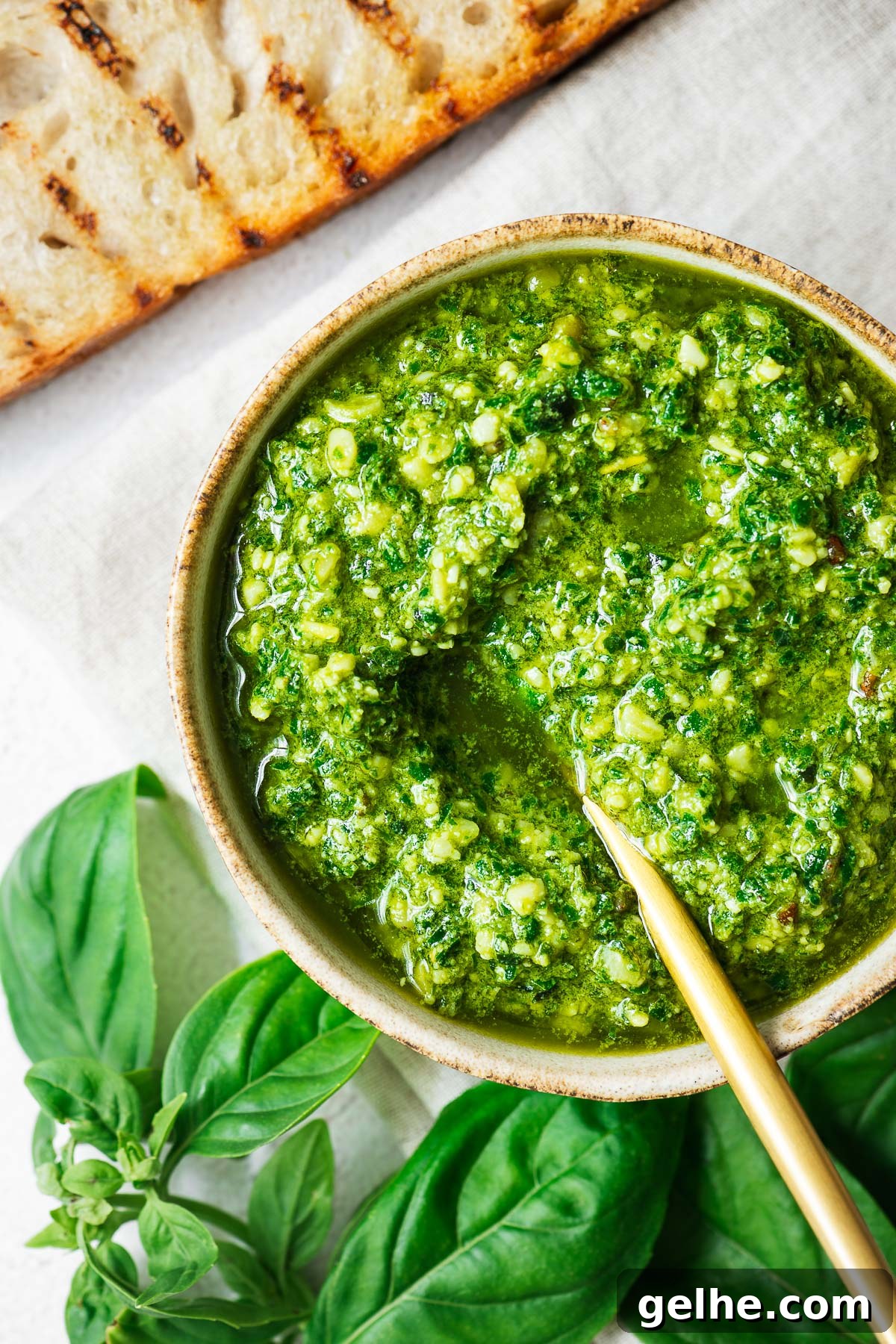 Top down view of a bowl of basil pesto surrounded by basil leaves with a slice of toasted sourdough in the upper left corner.