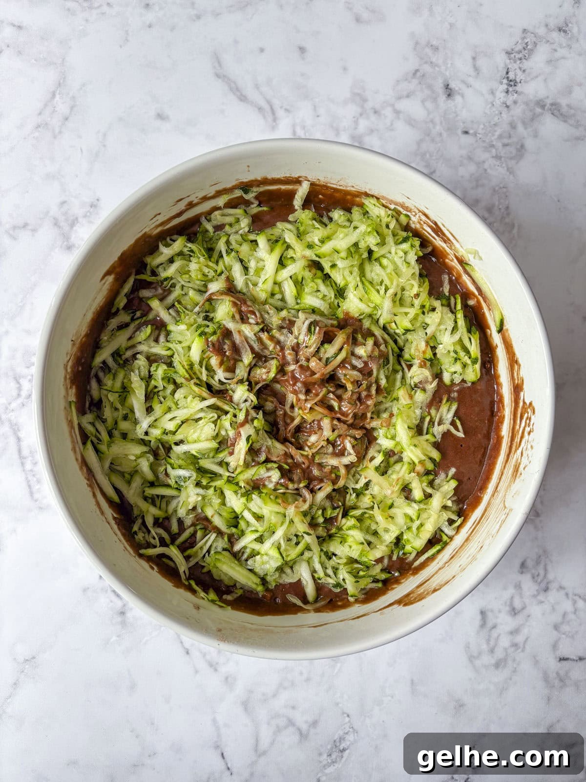 Overhead view of a mixing bowl with shredded zucchini being mixed into chocolate cake batter.