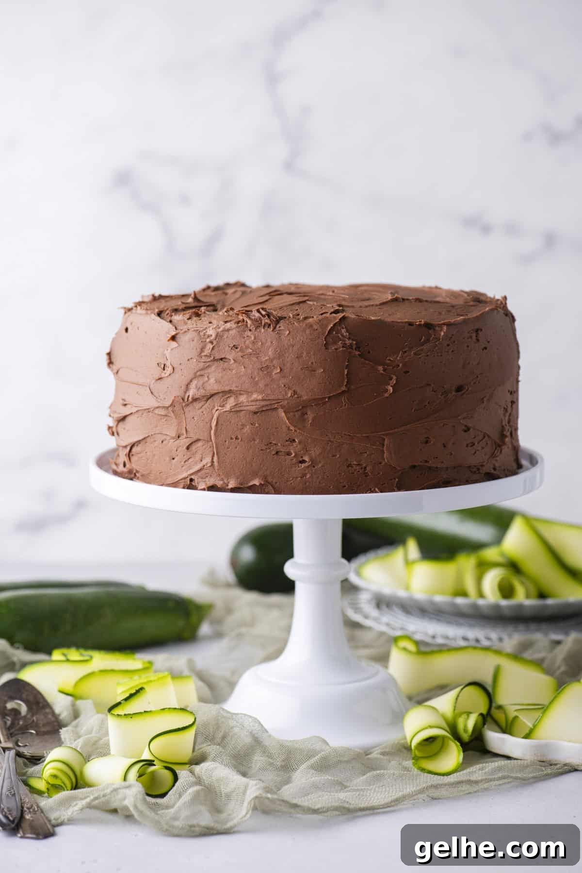 A chocolate frosted layer cake sits on a white cake stand, surrounded by green zucchini ribbons and whole zucchinis.