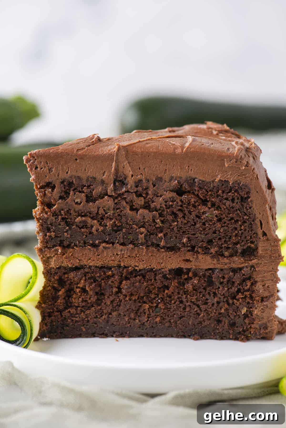 A close-up of a slice of chocolate layer cake with chocolate frosting on a white plate, garnished with a curl of yellow and green zucchini.