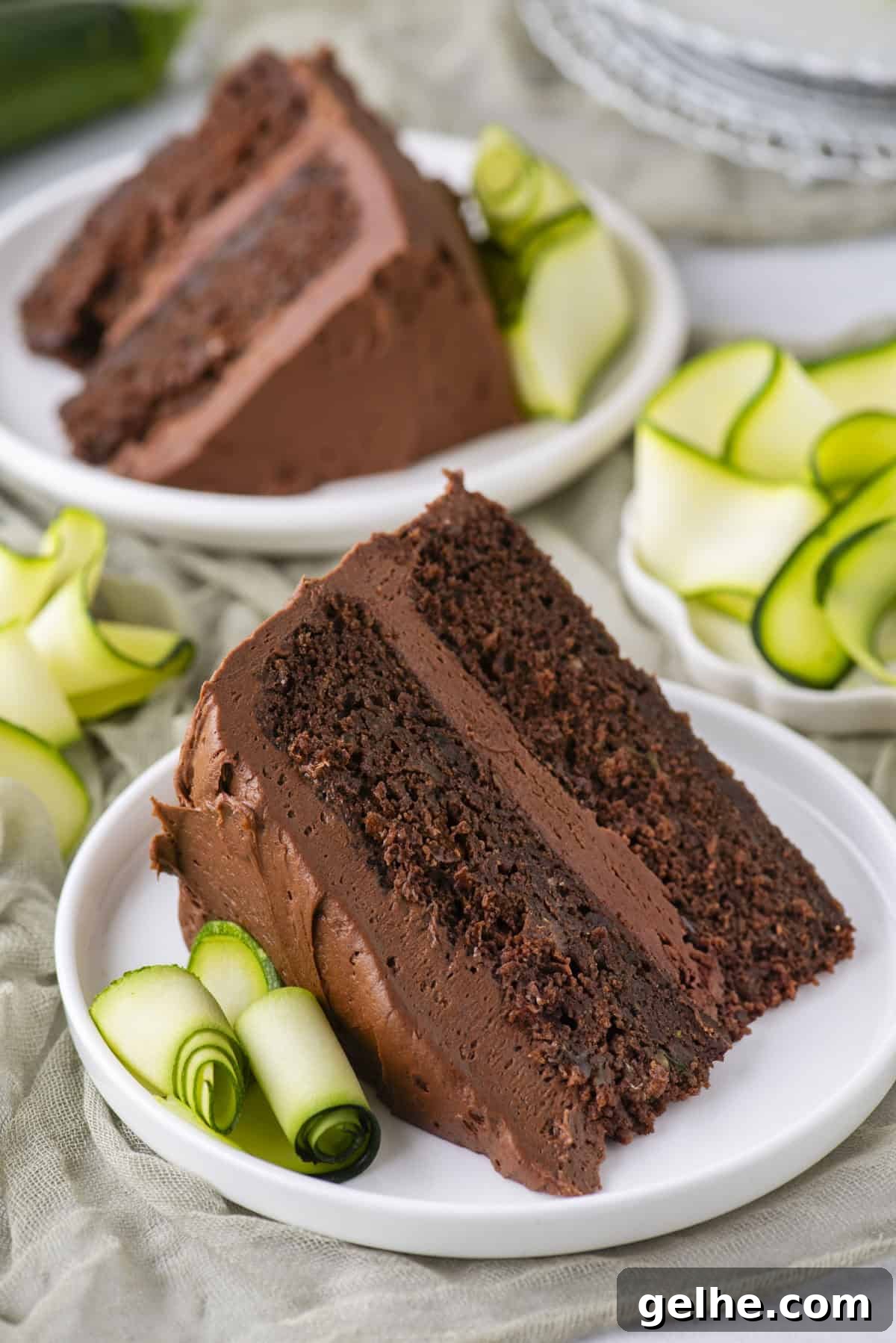 Two slices of rich chocolate cake with chocolate frosting are served on white plates, each garnished with thinly sliced, rolled zucchini ribbons.