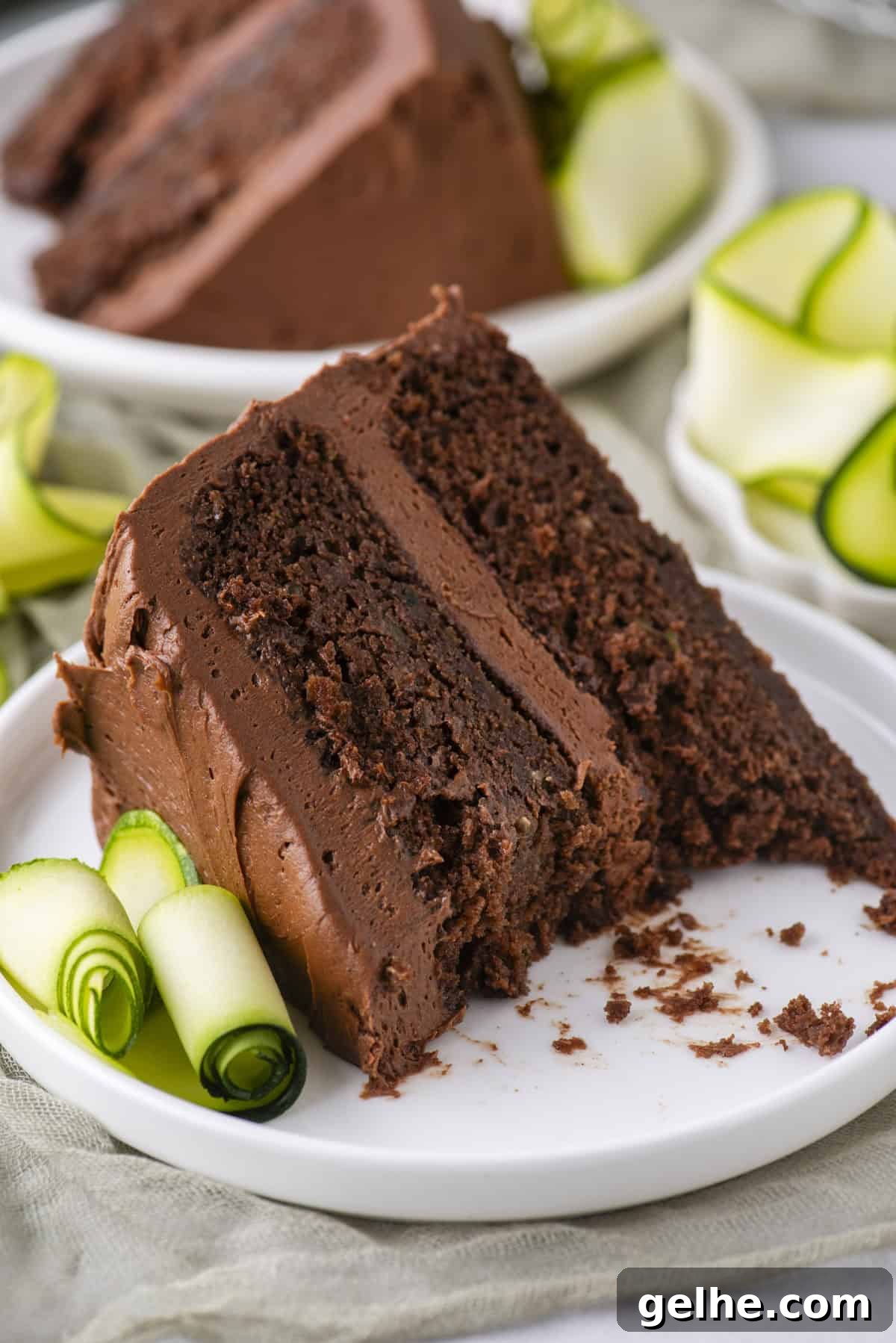 A slice of rich chocolate cake with chocolate frosting on a white plate, garnished with rolled slices of zucchini.