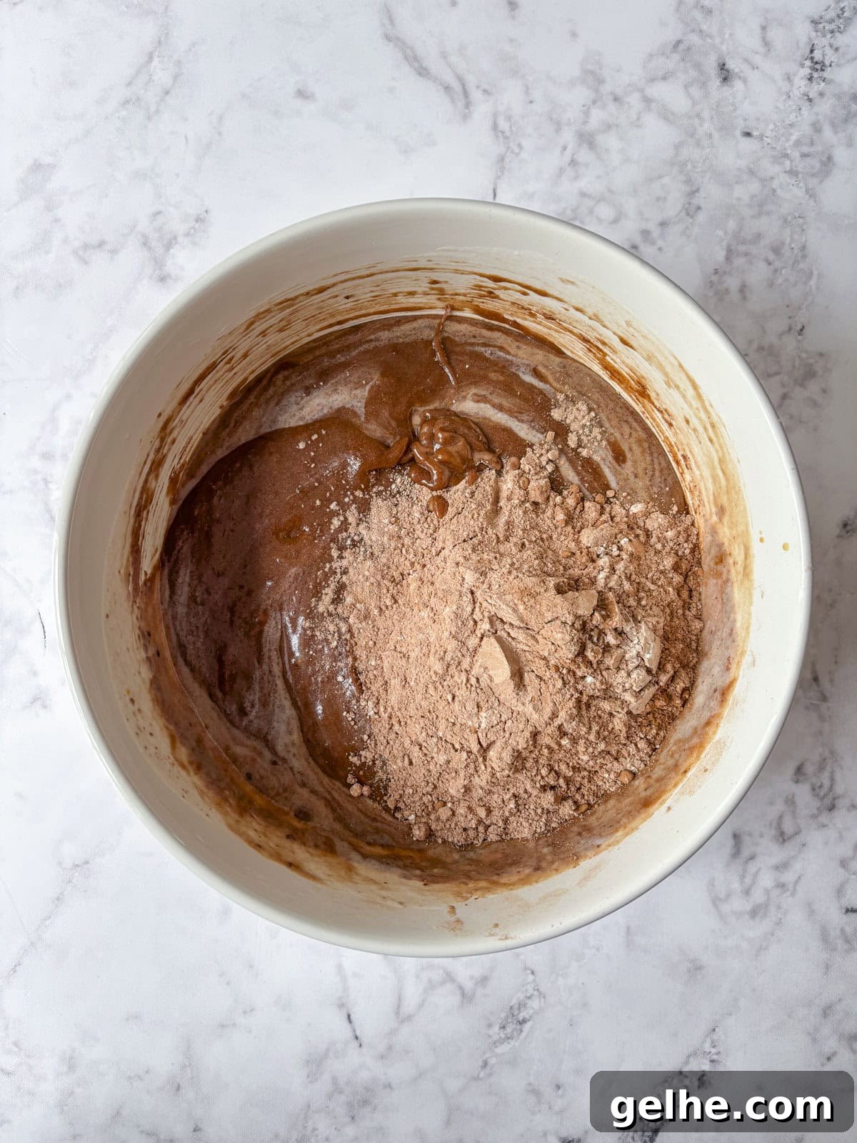 A white mixing bowl contains chocolate batter with some dry cocoa powder still visible on top.