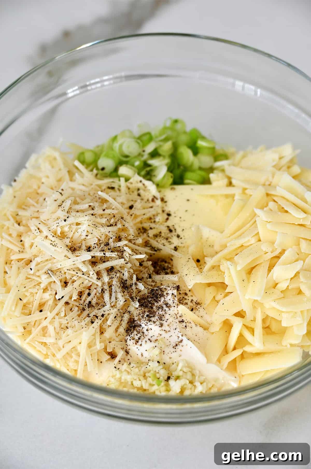 A glass bowl containing butter, shredded asiago and Monterey Jack cheese, sliced scallions, mayonnaise, salt and pepper.