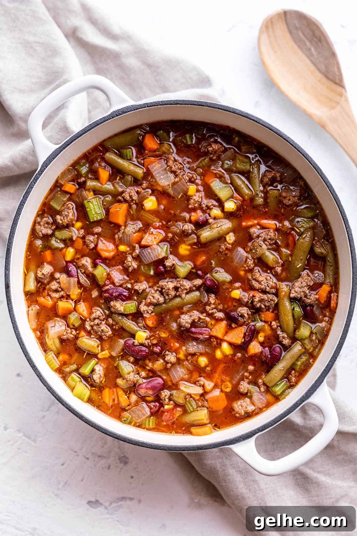 Hearty Ground Beef and Vegetable Soup Recipe 2 A white Dutch Oven full of a homemade soup with a wooden spoon on the side.