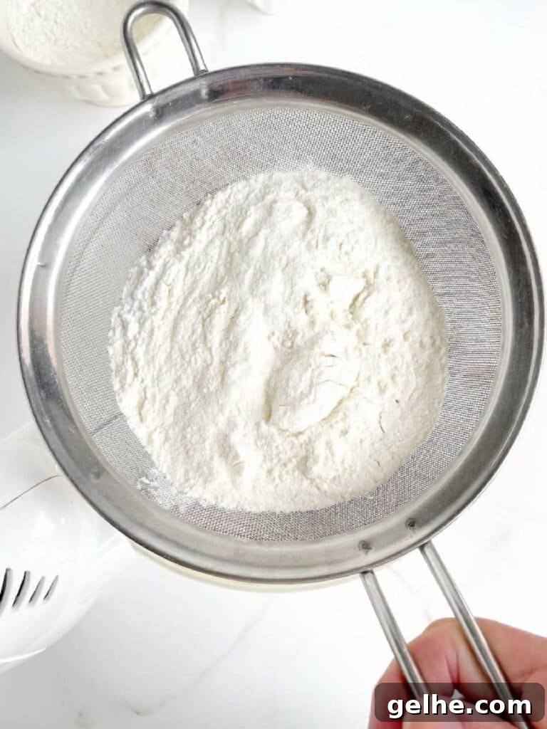 Sifting flour mixture over glass bowl with cake batter. White surface. Top view.