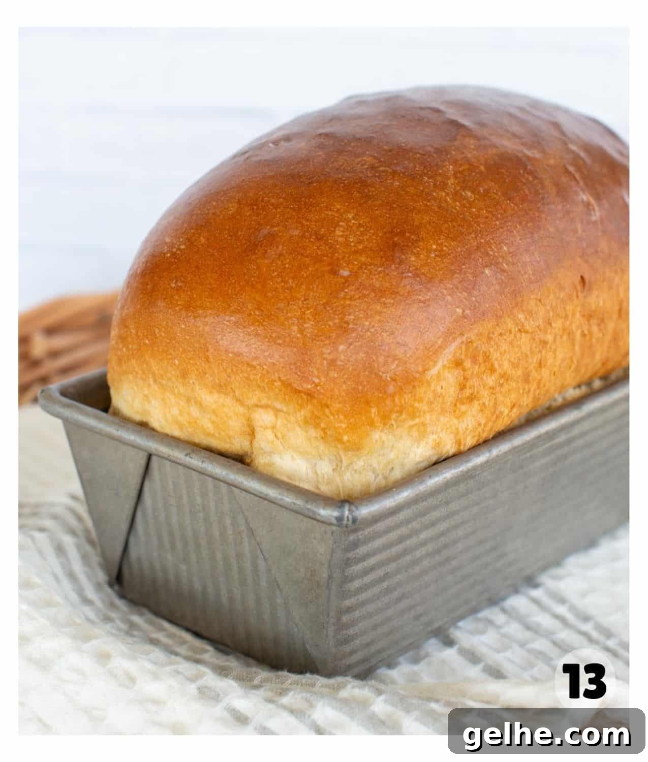 A golden brown loaf of white sandwich bread sits in a loaf pan on a kitchen towel.