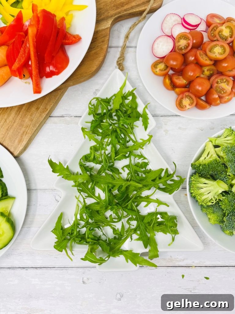 Image shows a christmas tree shaped plate with rocket scattered on it, next to a plate of broccoli, a plate of tomato and radish, a plate of peppers and a plate of cucumbers