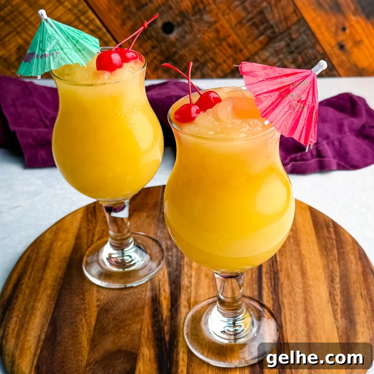 Two frozen Mai Tai slushies in curvy hurricane glasses, each garnished with cherries and colorful umbrellas, placed on a round wooden board.
