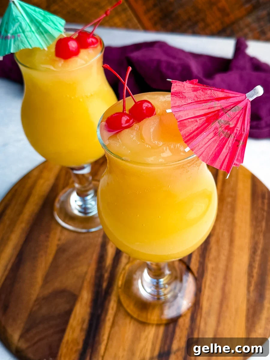 Two Mai Tai slushies garnished with maraschino cherries and paper umbrellas, displayed on a wooden board with a purple cloth behind them.