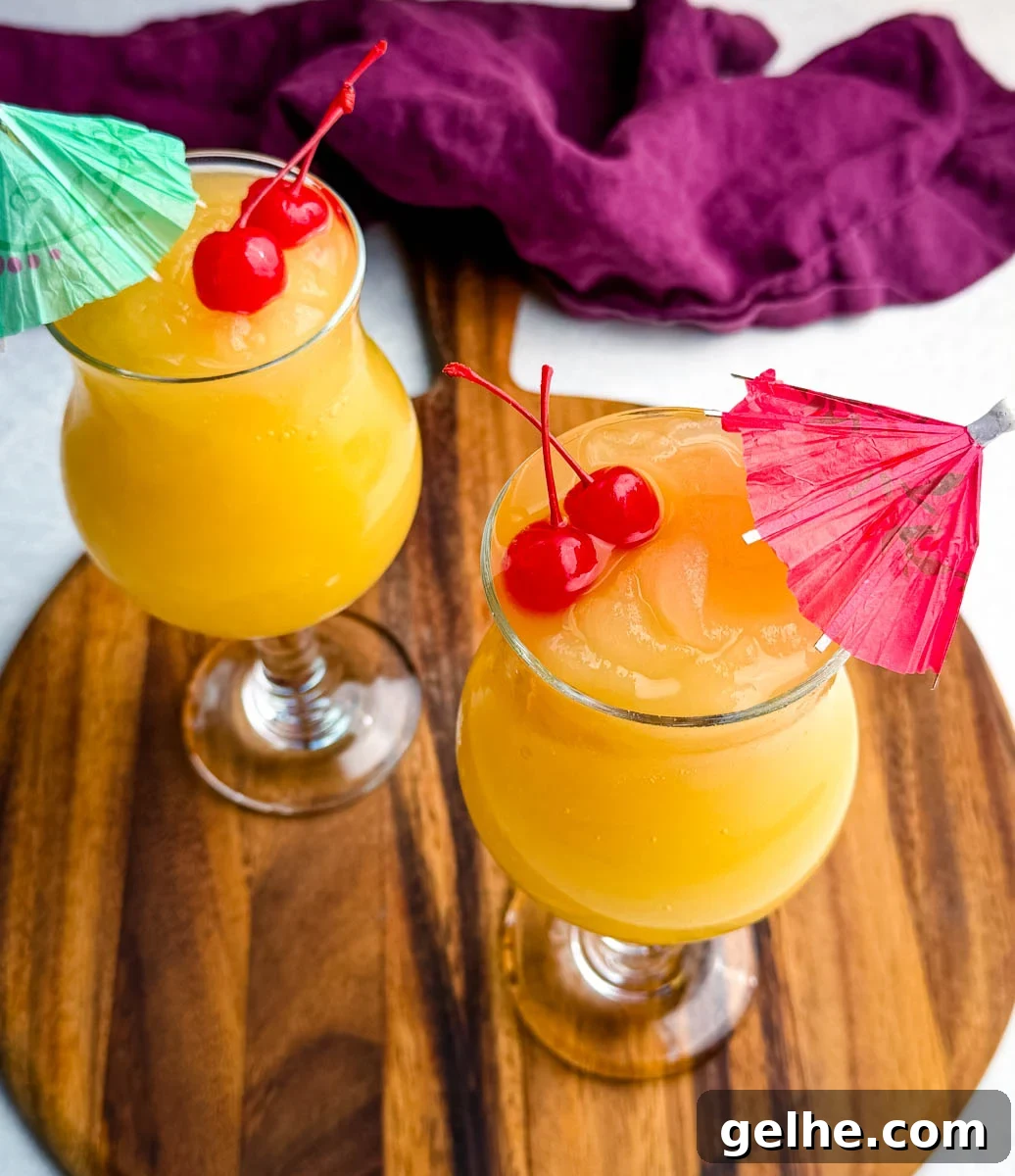 Two glasses of bright orange frozen Mai Tai slushies topped with maraschino cherries and paper drink umbrellas, sitting on a wooden board with a purple cloth in the background.