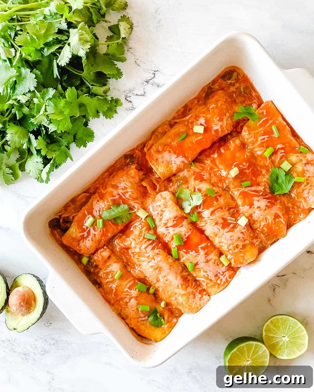 Slow Cooker Shredded Chicken Enchiladas Recipe 2 A baking dish full of chicken enchiladas is surrounded by cut limes, a halved avocado, and cilantro.