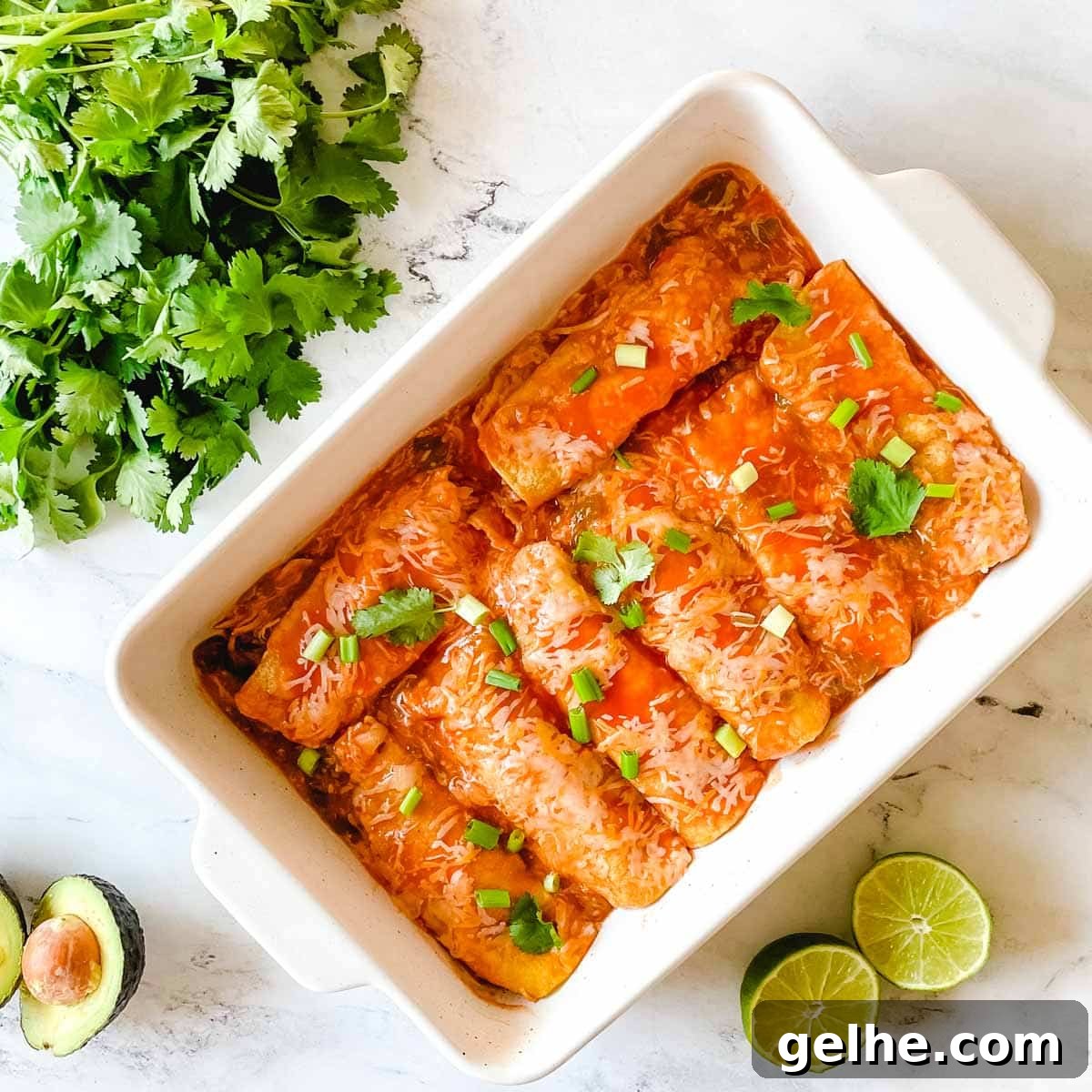 Slow Cooker Shredded Chicken Enchiladas Recipe 12 A baking dish full of chicken enchiladas is surrounded by cut limes, a halved avocado, and cilantro.