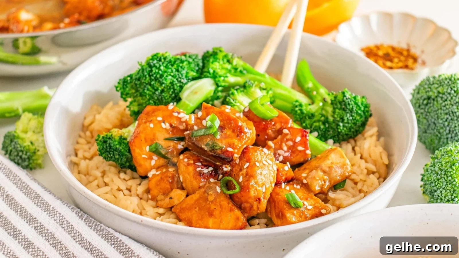 A bowl of brown rice topped with pieces of glazed chicken, broccoli florets, sesame seeds, and chopped green onions, with chopsticks resting on the side.