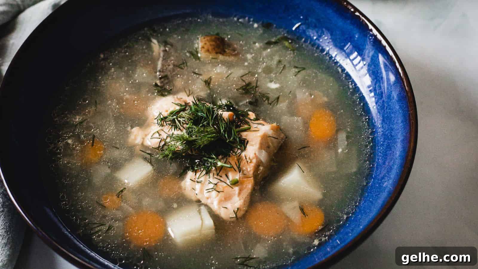 A blue bowl contains a soup with chunks of fish, carrots, and potatoes, garnished with fresh dill. The soup appears to have a clear broth base.