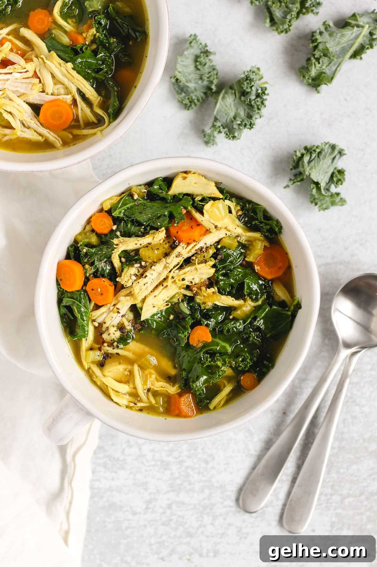 Chicken and kale soup in a white bowl with two soup spoons laid on the side.