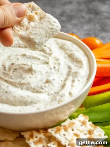 A hand dipping a cracker into a bowl of creamy pale herbed dip.