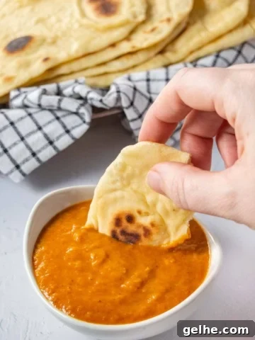 A hand dipping piece of naan bread into a small white bowl of orange sauce.