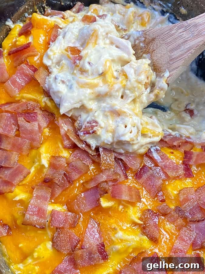 Close up of slow cooker shredded chicken covered in cheese and bacon with a wooden spoon lifting a serving out of the crock.