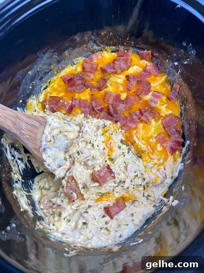Shredded chicken bacon ranch in a crockpot with cheddar cheese and a wooden spoon stirring it together.
