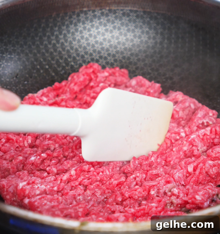 Breaking up raw ground beef in a nonstick skillet with a white spatula to begin cooking honey garlic beef. The beef is starting to brown and cook evenly.