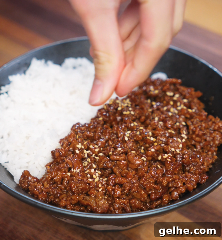 Garnishing a honey garlic beef rice bowl with toasted sesame seeds. The glossy beef is served next to a bed of white rice in a black bowl.