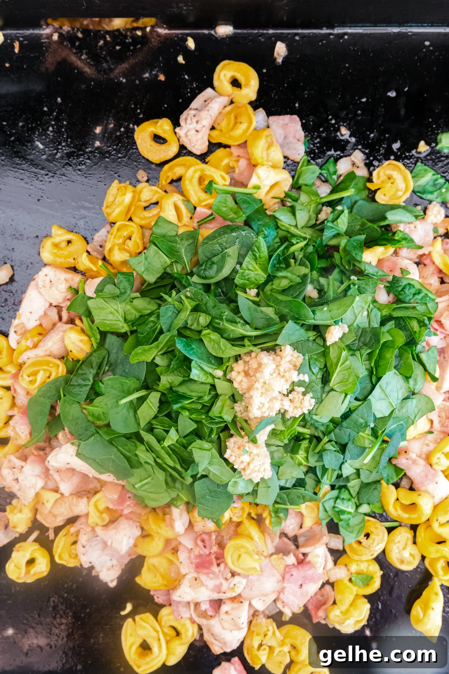Chicken, bacon, tortellini, fresh spinach, and minced garlic cooking together on a flat-top griddle.