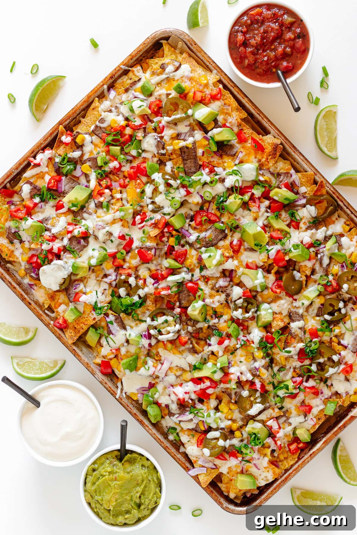 Loaded Steak Nachos Recipe with Melted Cheese 2 Sheet pan of steak nachos surrounded by lime wedges and small bowls of salsa, sour cream and guacamole.