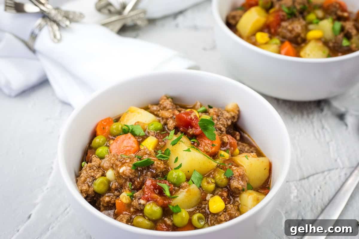 Two bowls of Homestyle hamburger stew with chunks of potato, carrots, peas, corn, and parsley garnish.