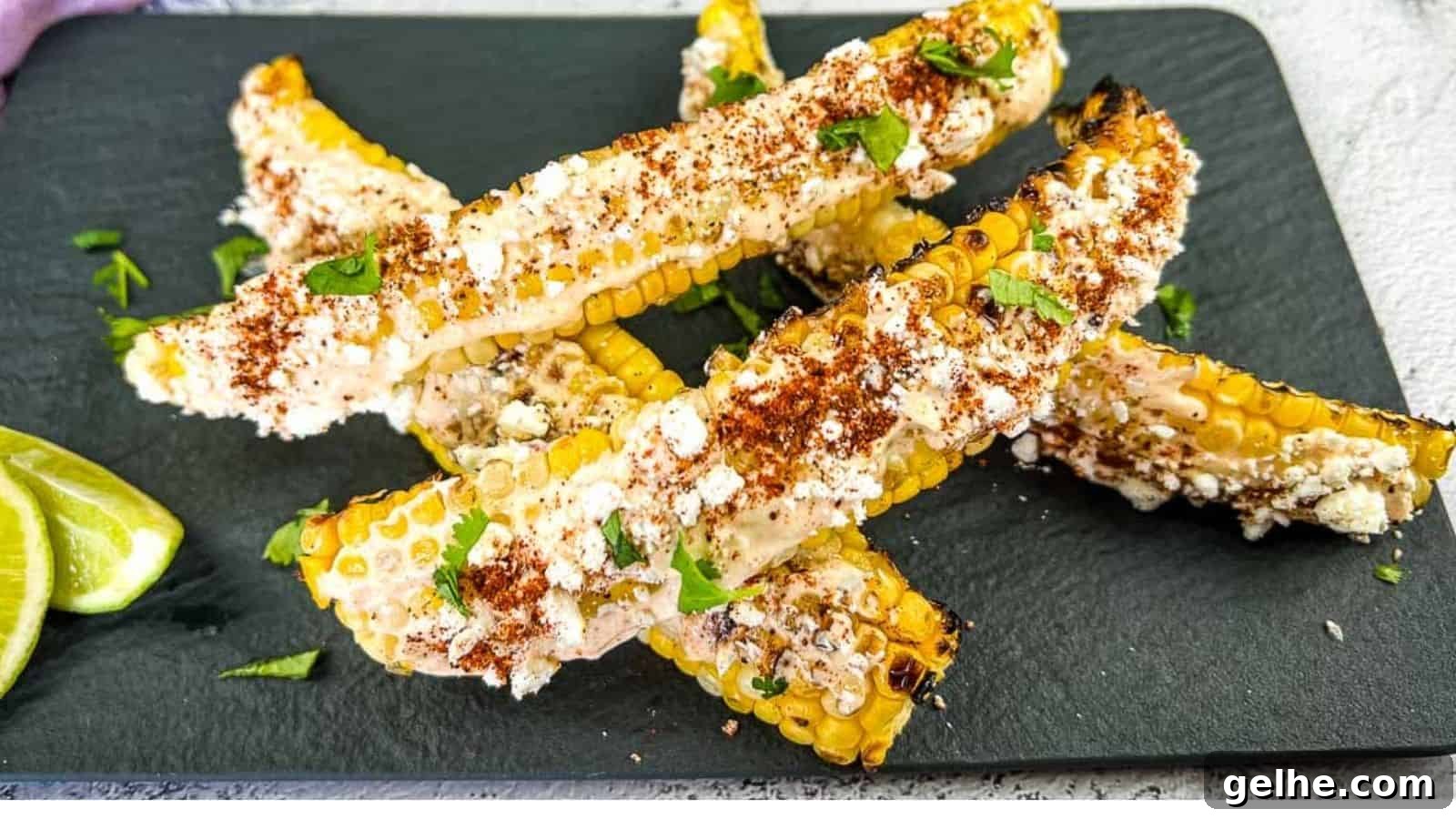 Grilled corn on the cob pieces topped with cheese, chili powder, and cilantro, served on a black slate plate with lime wedges on the side.